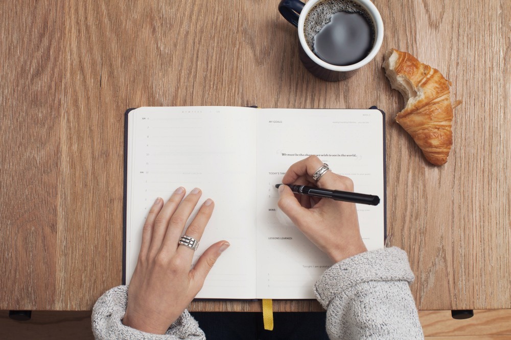 a hand writing on a notebook. Coffee and a half-eaten croissant on the side