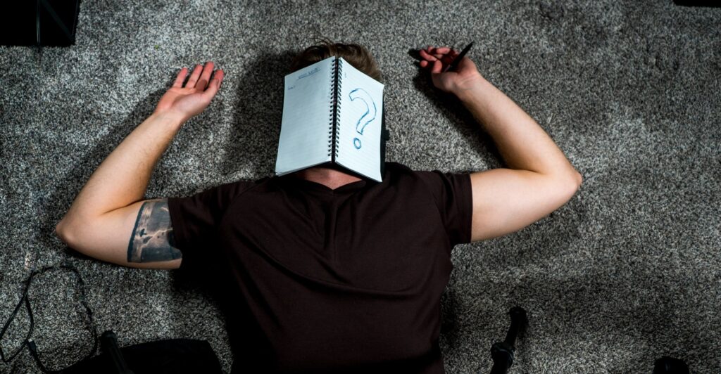 A man lying on the floor with a notebook on his head. The notebook has a question mark drawn on it